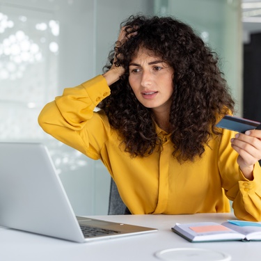 A woman in a bright yellow shirt anxiously places a hand to her head as she looks at her computer and holds a credit card.