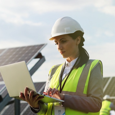 A woman engineer wearing a white hard hat stands outside next to solar panels while using a laptop.