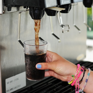 A close-up view shows a person holding a small, clear, plastic cup under a drink dispenser. Brown liquid is filling the cup.