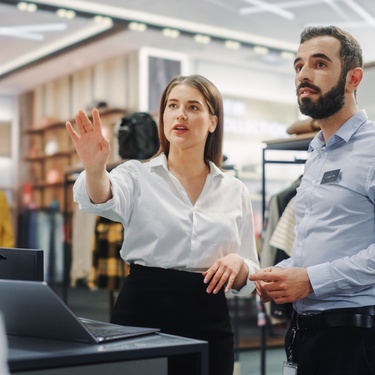 Two retail managers are planning something in the clothing store. They're looking at a mannequin with their laptops.