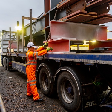A worker in orange safety gear secures large steel beams on a blue flatbed truck at an industrial site.