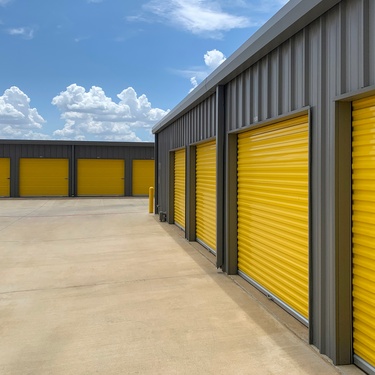 The exterior of a gray storage facility with a large concrete driveway and a yellow garage door on each unit.