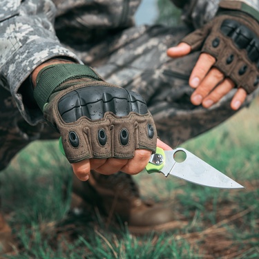 A man is crouching and holding a small knife in the forest. He wears military gear, including gloves and boots.