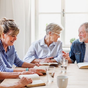 A group of older adults sits at a table in a sunlit room. Each has an open book, and one man gestures as he speaks.
