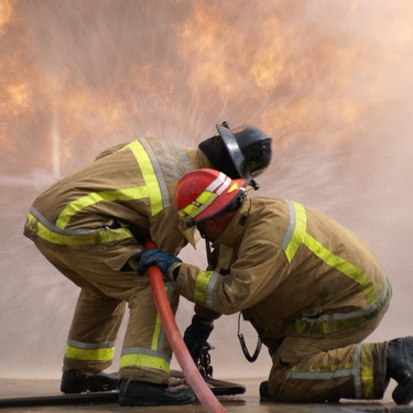 Two wildland firefighters wearing protective equipment stand close to each other while holding up a water hose.
