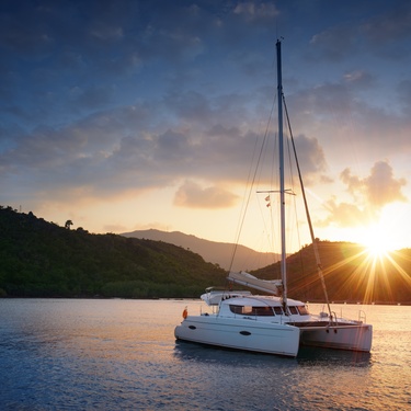 A white catamaran with a tall sail floating on a smooth body of water surrounded by green hills at sunset.