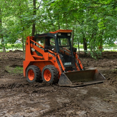An orange skid steer loader with a front bucket moving dirt in a wooded area, clearing muddy ground among green trees.