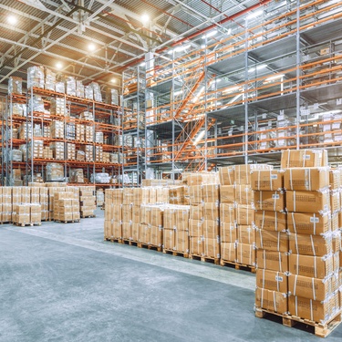 A spacious warehouse with tall shelves stocked with boxes and additional cardboard boxes stacked on pallets on the floor.