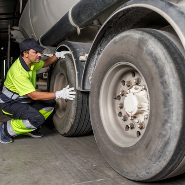 A man wearing a blue and yellow safety outfit and blue baseball cap inspects the tire of a trailer.