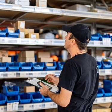 A warehouse worker with a clipboard is checking the warehouse's inventory. He is looking at shelves of small containers.