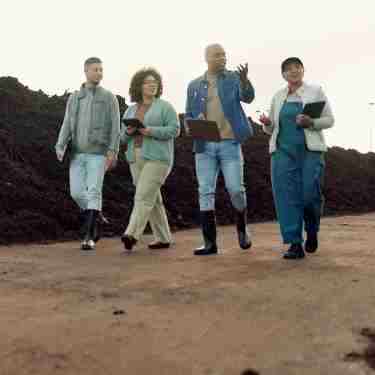 A team of four walks and examines a piece of land. There is a large pile of dirt that rises up behind them.