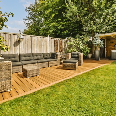 An outdoor patio with weather-resistant black sofa chairs on top of wooden planks, surrounded by various greenery.