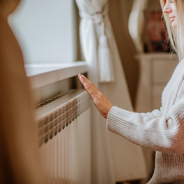 Young woman in a long beige winter jumper stands at home near a radiator, staying cosy on a cold day.