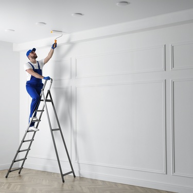 A man wearing blue overalls is standing on a ladder while painting the ceiling. The walls are white and have a trim.