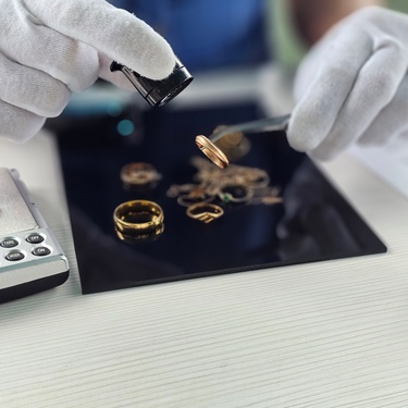 A person wearing white gloves inspects gold rings and jewelry with a loupe and tweezers beside a digital scale.