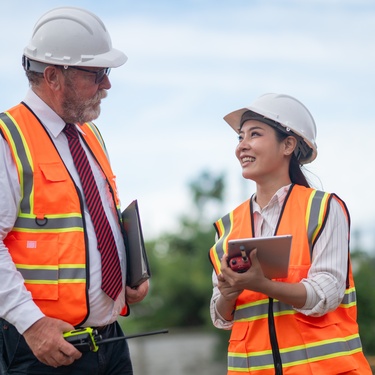 Two people outside wearing reflective vests and hard hats. One of them holds a tablet and the other has a binder.