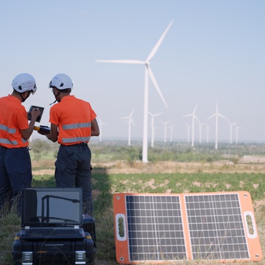 Two men in reflective orange shirts review data on a tablet while standing near wind turbines and a solar panel.