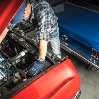A mechanic wearing a plaid shirt looks under the open hood of a classic red muscle car in a restoration workshop.
