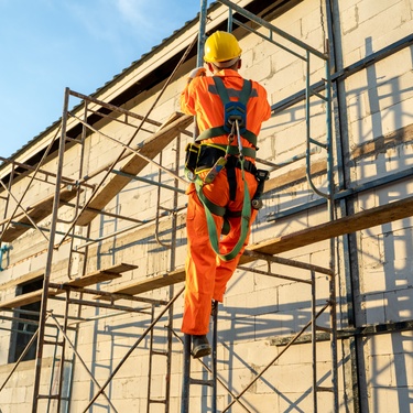 A construction worker wears a high-vis orange suit with a yellow helmet as they climb the scaffolding.