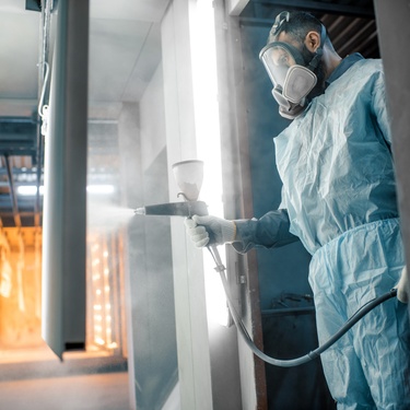 A worker in protective gear applies a coating to a metal surface inside a large industrial factory setting.