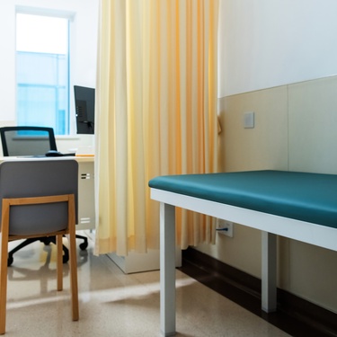 An empty doctor’s office with a long green bench and a small desk with two chairs. A computer is on the desk.