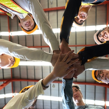 Seven workers in hard hats and long sleeves stacking their hands together in a circle with their palms flat and facing down.