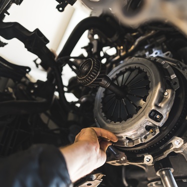 A mechanic's hand reaches toward a vehicle's clutch. The hand is reaching in from the car's underside.