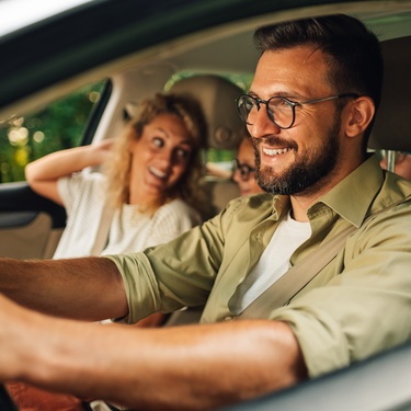 Two parents smiling and driving in a car during the daytime. Their two kids sit in the back seat of the vehicle.