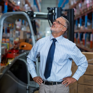 A professionally dressed person looking upward while standing in a warehouse surrounded by large shelves and boxes.