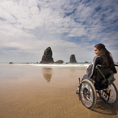A woman sits in her wheelchair on a sandy, wet beach. She smiles to herself as she looks out at the landscape.