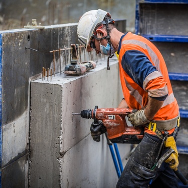 A man wearing a reflective vest, a white hard hat, a mask, and safety glasses drills into a concrete wall.