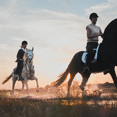 Two women, both wearing safety helmets, riding horses across a muddy field beneath a partly cloudy sunset sky.