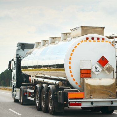 A tanker truck is on an empty highway in the right lane. The tanker truck features symbols for hazardous materials.