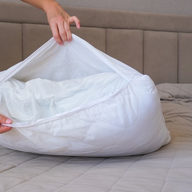 Two hands from out of frame delicately remove the pillow cover from a pillow resting on a bed with beige bedding.