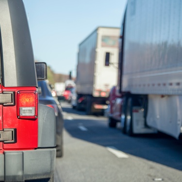 A red truck stuck beside a semi-trailer and behind a small black car in a traffic jam on a clear day.
