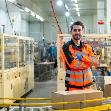 A man in a work uniform stands by a conveyor belt moving two cardboard boxes. There is other machinery around him.