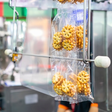 A close-up of sandwich cookies wrapped in a clear pouch hanging from a packaging machine in a factory.