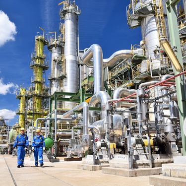 The exterior of an industrial chemical plant with a lot of pipes and machinery. Two workers in blue gear walk outside.
