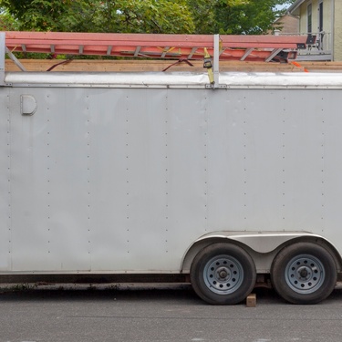 A white enclosed dual-axle work trailer parked on a residential street with ladders secured on the top rack.