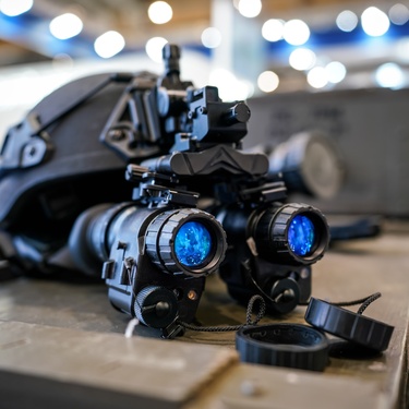 A night vision helmet sitting on a tabletop. The helmet is dark gray with a blue reflective lens on the goggles.