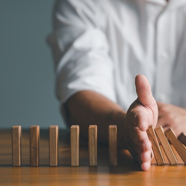 A hand blocks falling wooden dominoes on a table, stopping the chain while untouched pieces remain upright beside it.