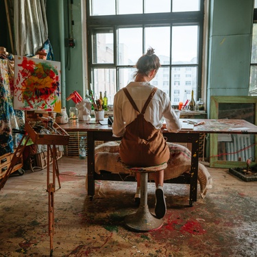 A woman working at a table in an art studio. The space is messy but organized and covered in paint and crafting tools.