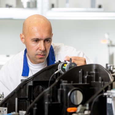 A man in a white lab coat stands behind a part. He uses various equipment to take measurements of the component.