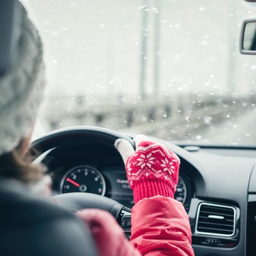 A person driving their car on a snowy day. The driver is wearing pink gloves, a pink jacket, and a gray-and-white hat.