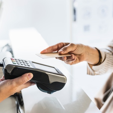 Two people at a reception desk. One is holding a card reader, and the other is moving their credit card towards it to use it.