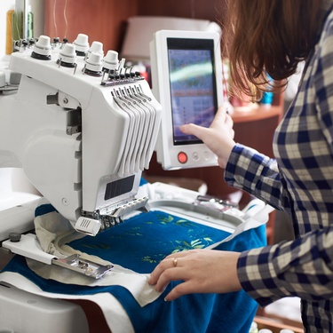 A woman in a flannel shirt uses the screen of an embroidery machine to set a design to stitch into blue fabric.