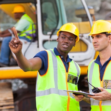 Two employees talking outside on a construction site. A third employee is sitting in a crane behind them.