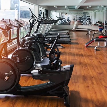 A room with hardwood floors, a mirror along one wall, and large windows full of exercise equipment.