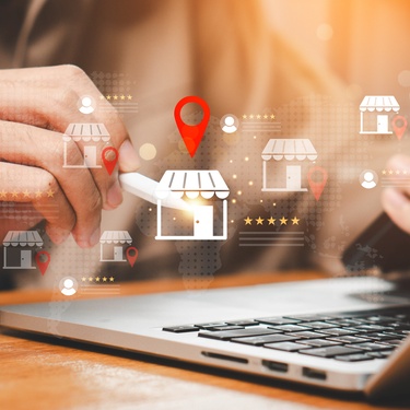 Someone on their laptop while holding their phone and a pencil. The pencil is pointing at a storefront graphic.