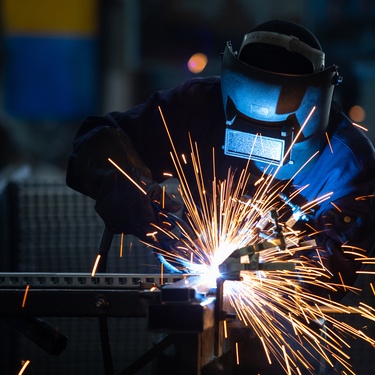 Someone wearing a mask and other protective gear as they weld a metal piece. Sparks are flying as they work.